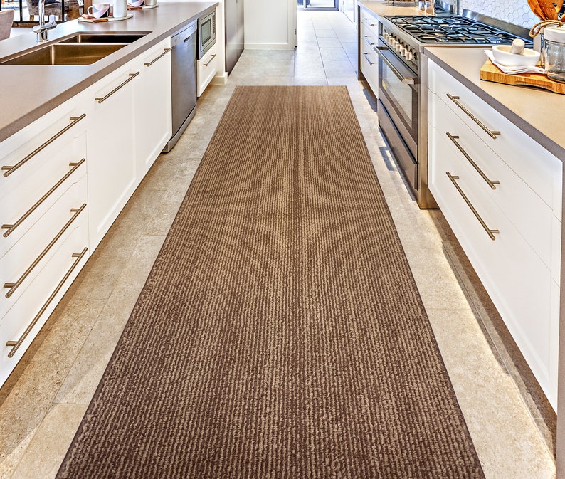 May include: A brown and beige striped kitchen runner rug with a subtle sheen. The rug is placed in a kitchen with white cabinets and a tiled floor.