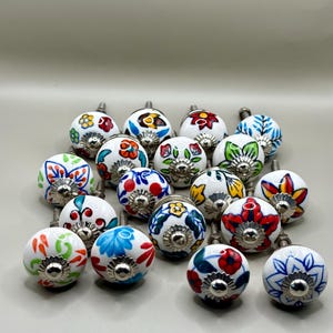 Peut inclure: Ensemble de boutons de meubles en céramique décoratifs. Chaque bouton blanc est orné d'un motif floral peint à la main dans des couleurs variées : rouge, bleu, jaune et vert. Les boutons sont équipés d'une fixation métallique argentée.