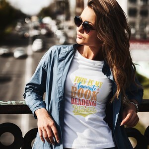 May include: A woman wearing a white t-shirt with the text "This is my favorite book reading blanket" in colorful letters. She is wearing a blue denim jacket and blue jeans.