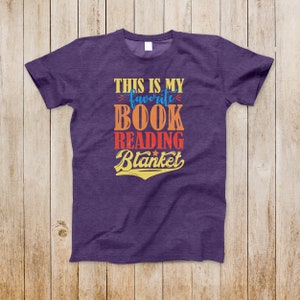 May include: Purple t-shirt with the text "This is my favorite book reading blanket" in a vintage style font with a star.