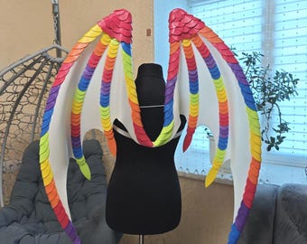 May include: Rainbow-colored dragon wings with a white membrane, displayed on a black mannequin. The wings feature a layered design with red, orange, yellow, green, blue, and purple scales. The wings are designed for costume or cosplay.