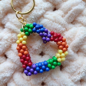 May include: A rainbow-colored beaded heart keychain with a gold-colored ring. The heart is made of small, round beads in a gradient of colors, including red, orange, yellow, green, blue, and purple. The keychain is on a soft, light pink textured background.