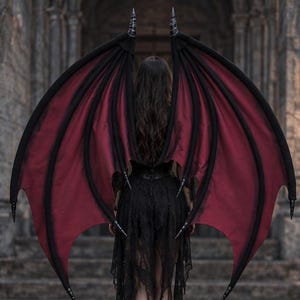 May include: Large, articulated bat-like wings featuring a deep red fabric membrane stretched across a black structural frame. The wings have pointed, horn-like tips at the top and bottom, with a dark, skeletal design that extends outwards.