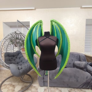 K&ouml;nnte beinhalten: Gr&uuml;n-schwarze Drachenfl&uuml;gel-Kost&uuml;m mit schuppiger Textur. Die Fl&uuml;gel sind an einem schwarzen Mannequin befestigt.