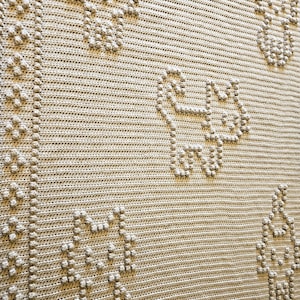 Könnte beinhalten: Nahaufnahme einer beige gehäkelten Decke mit einem sich wiederholenden Muster aus stilisierten Katzenfiguren und floralen Designs. Die Decke weist ein strukturiertes, erhabenes Stichmuster auf, das eine taktile und visuell interessante Oberfläche schafft. Das Gesamtdesign ist schlicht und elegant.