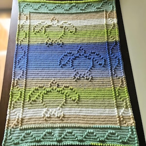 May include: A crocheted blanket with a green, blue, and beige color scheme. The blanket features a repeating pattern of small circles.