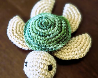 May include: A handmade crochet turtle toy. The turtle features a green and white shell with a spiral design, four cream-colored flippers, and a cream-colored head with black button eyes. The toy is made of soft yarn.