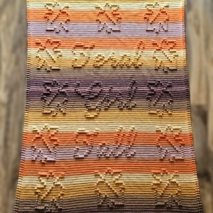 May include: A handmade, rectangular blanket with a gradient color scheme of purple, orange, and yellow. The blanket features the words "Be Still" and decorative floral patterns, all in a raised, textured design.