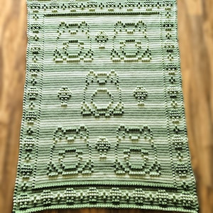 Puede incluir: Una manta de ganchillo hecha a mano, verde y blanca, con un patrón repetido de diseños de ranas. La manta tiene un patrón de puntada en relieve texturizado y un borde decorativo. El patrón de rana está hecho con un punto de bobble.