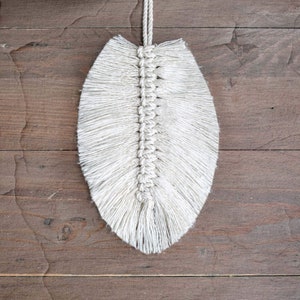 May include: A white macrame feather hanging from a wooden surface. The feather is made of knotted string and has a fringe at the bottom.