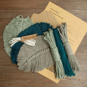 May include: A macrame feather kit with instructions. The kit includes three skeins of yarn in teal, sage green, and grey. The yarn is wrapped around a tag that says "Rocco & Arlo".