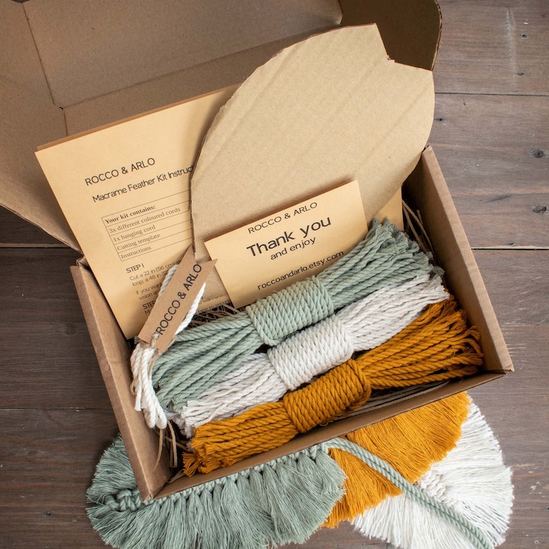 May include: A macrame feather kit in a cardboard box. The kit includes three different colored cords, a hanging cord, a cutting template, and instructions. The box also includes a thank you note from Rocco & Arlo.