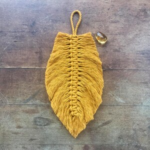 May include: A mustard yellow macrame feather wall hanging. The feather is made with a knotted string technique and has a fringe at the bottom.