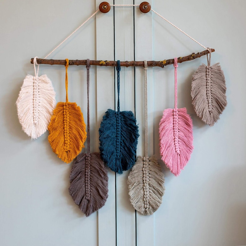 May include: A wall hanging made of macrame feathers in various colors, including white, yellow, blue, gray, and pink. The feathers are attached to a branch and hang from a string.