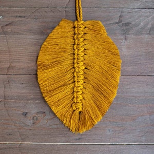 May include: A mustard yellow macrame feather wall hanging with a long, thin shape. The feather is made of knotted yarn and has a fringe at the bottom.