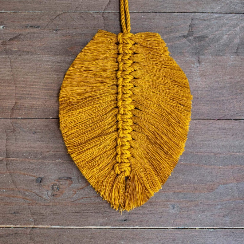 May include: A mustard yellow macrame feather wall hanging with a long, thin shape. The feather is made of knotted yarn and has a fringe at the bottom.