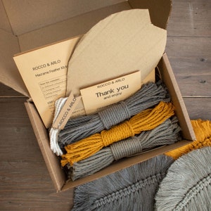 May include: A macrame feather kit in a cardboard box. The kit includes three balls of cord, one grey and two yellow, a cardboard template, and instructions. The box also includes a thank you note that reads "Thank you and enjoy".