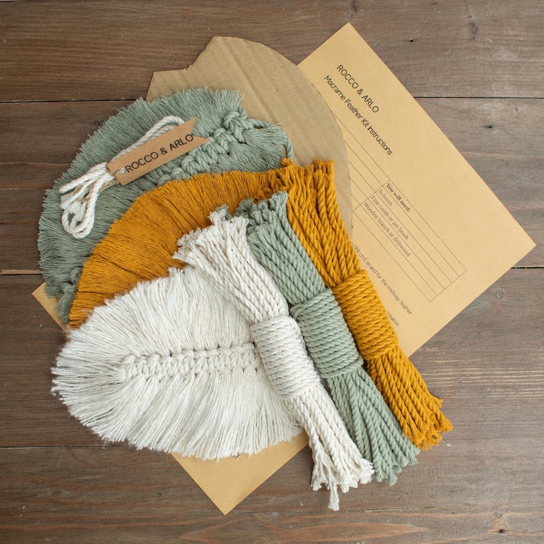 May include: A macrame feather kit with instructions, including three skeins of yarn in mustard yellow, sage green, and natural white. The kit also includes a wooden dowel and a comb or brush for finishing the macrame feather.