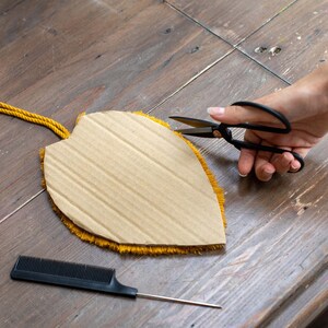 May include: A hand holds black scissors cutting a yellow fringe around a cardboard shape. The shape is a leaf with a brown cardboard center. A black comb and a thin metal needle are on the wooden surface.