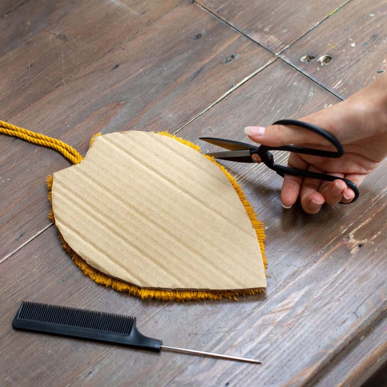 May include: A hand holds black scissors cutting a yellow fringe around a cardboard shape. The shape is a leaf with a brown cardboard center. A black comb and a thin metal needle are on the wooden surface.