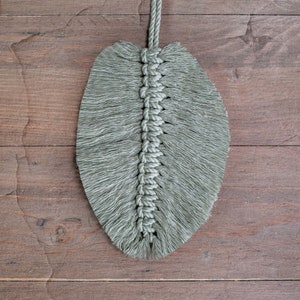May include: A green macrame leaf wall hanging with a fringe border. The leaf is made with a knotted rope and has a long cord for hanging.