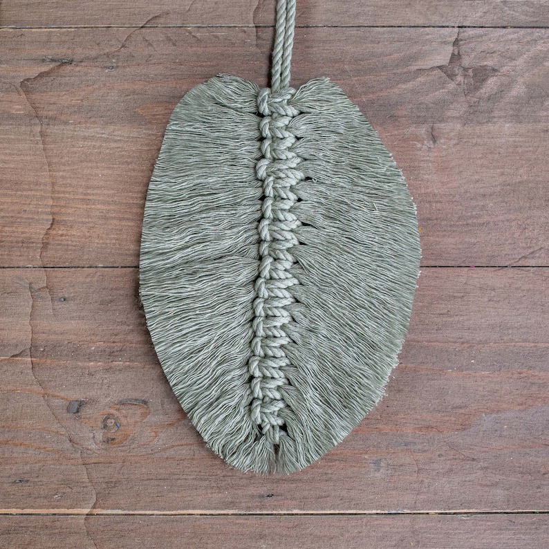 May include: A green macrame leaf wall hanging with a fringe border. The leaf is made with a knotted rope and has a long cord for hanging.