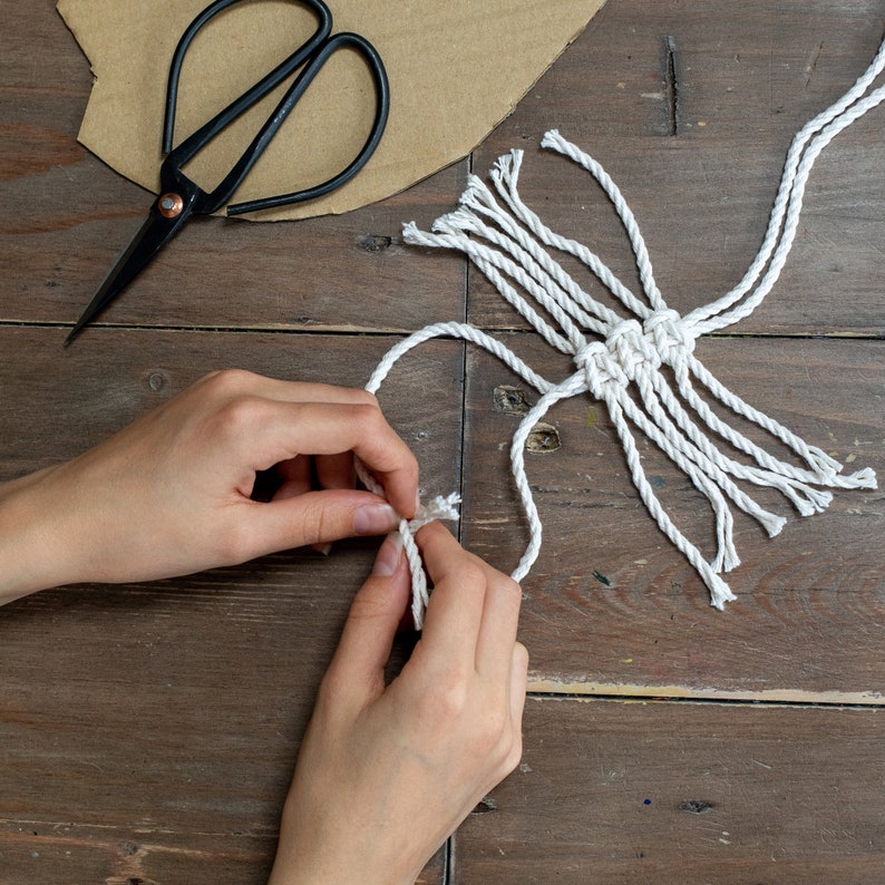 May include: A person's hands are tying a white macrame knot. The knot is made with a thick white cord and has a fringe on the bottom. The knot is on a wooden surface.
