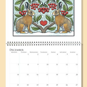Op de afbeelding: Een decemberkalenderpagina met een illustratie van twee bruine hazen onder een gestileerde boom met rode bessen en een hart. De kalender is spiraalgebonden en toont data en feestdagen.