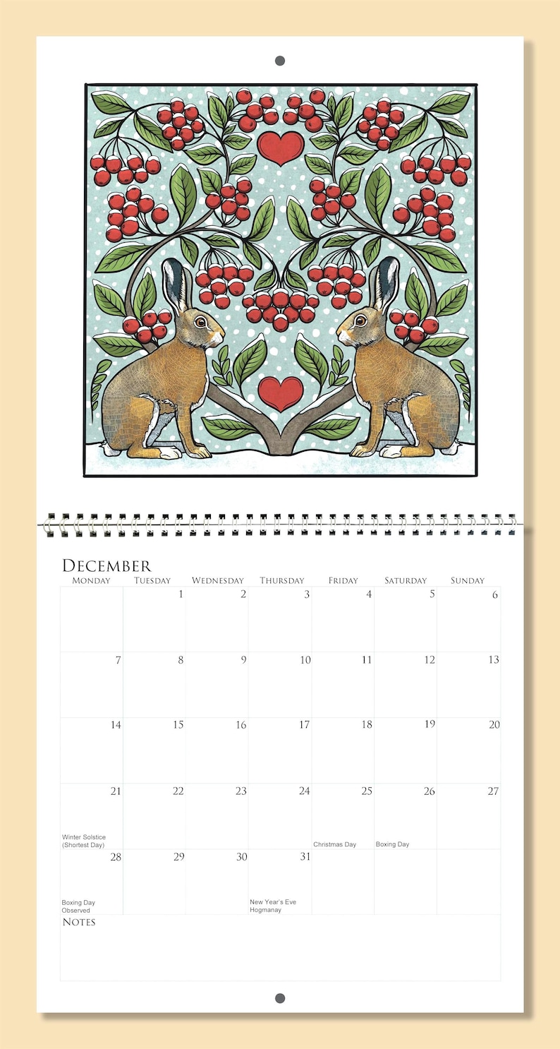 Op de afbeelding: Een decemberkalenderpagina met een illustratie van twee bruine hazen onder een gestileerde boom met rode bessen en een hart. De kalender is spiraalgebonden en toont data en feestdagen.