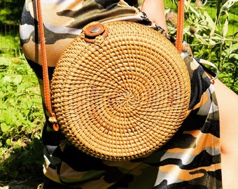 May include: A round, woven rattan handbag with a brown leather strap and accents. The bag is light brown and has a circular design. The strap is attached to the bag with a small, circular brown leather piece.