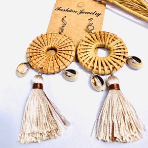 May include: A pair of woven rattan earrings with white tassel accents and small seashells hanging from the bottom. The earrings are attached to silver hooks. The card behind the earrings reads "Fashion Jewelry".