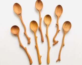 Puede incluir: Siete cucharas de madera con un diseño natural y orgánico. Cada cuchara tiene un cuenco redondeado y un mango con forma de rama de árbol. Las cucharas son de color marrón claro cálido y están dispuestas sobre una superficie blanca.