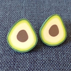 Può includere: Due orecchini a forma di avocado. Ogni orecchino ha un bordo esterno verde, una sezione interna bianca e un seme marrone. Su uno sfondo blu strutturato.
