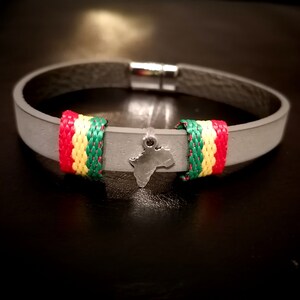 Könnte beinhalten: Graues Lederarmband mit einem silbernen Anhänger des afrikanischen Kontinents und einem rot-gelb-grünen Streifenband.