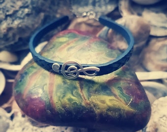 Peut inclure: Un bracelet en cuir noir avec un charme en argent en forme de clé de sol. Le bracelet est sur un rocher coloré.