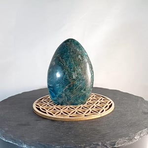 Op de afbeelding: Een gepolijste, eivormige steen in teal met donkere adering, rustend op een ronde, lasergesneden houten standaard. De standaard heeft een geometrisch patroon. De steen is ongeveer 13 cm hoog.