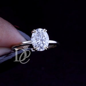 May include: A silver-toned ring featuring a single oval-cut clear gemstone set in a four-prong setting. The band is thin and polished, and a small logo with the letters DE and a diamond icon is visible on the metal surface near the setting.