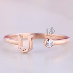 Puede incluir: Anillo de oro rosa con la letra "U" y una pequeña gema transparente. El anillo tiene un diseño delicado y abierto. El fondo está borroso, con tonos rosas y blancos.