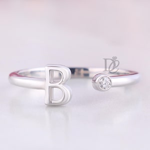 May include: A silver-toned open ring featuring a capital letter B on one end and a single small, round, clear stone set in a bezel on the opposite end. The band has a smooth, polished finish and a small logo mark is visible near the stone.