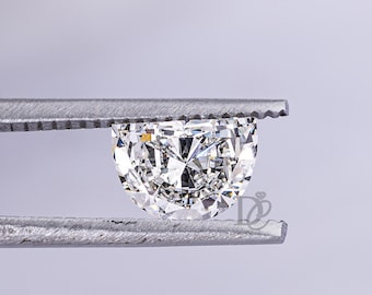 May include: A loose, brilliant-cut, semi-circular diamond held by silver-toned metal tweezers. The gemstone features a clear, faceted surface that reflects light, displaying a high level of clarity and sparkle against a light background.