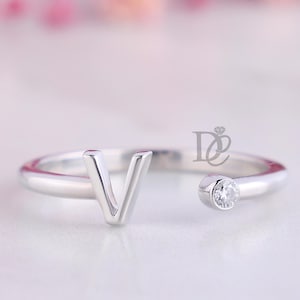 May include: A silver-toned open ring featuring a capital letter V on one end and a single round clear stone set in a bezel on the other end. The ring has a smooth, polished finish and includes a small logo mark with the letters DE.