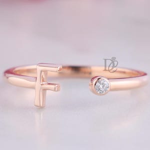May include: A rose gold open ring featuring a capital letter F on one end and a single round clear stone set in a bezel on the opposite end. The band is smooth and polished, showcasing a minimalist design with a small logo mark near the stone.