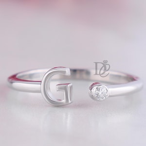 Puede incluir: Un anillo abierto de tono plateado que presenta una letra G mayúscula en un extremo y una pequeña piedra redonda y transparente engastada en el otro. El anillo tiene un acabado liso y pulido e incluye un pequeño logotipo con las letras DE.