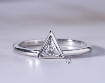 May include: A silver-toned ring featuring a single triangular-cut clear gemstone set in a matching triangular metal frame. The band is smooth and polished, showcasing a clean, geometric design with a small logo mark visible near the base of the setting.