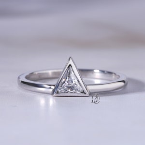 May include: A silver-toned ring featuring a single triangular-cut clear gemstone set in a matching triangular metal frame. The band is smooth and polished, showcasing a clean, geometric design with a small logo mark visible near the base of the setting.