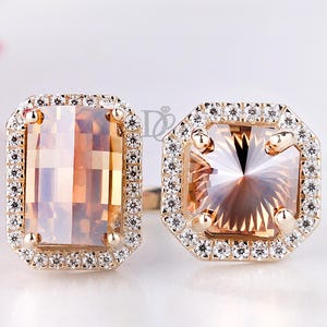May include: Two gold rings with large, rectangular and square, peach-colored gemstones. Each gemstone is surrounded by a halo of small, clear stones. The rings are set against a white background.