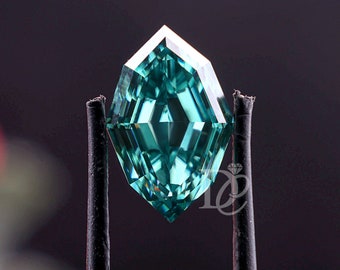 May include: A loose, faceted gemstone with a teal-green hue and a shield-like geometric cut. The stone features sharp, symmetrical facets that reflect light, and it is held by a pair of metal tweezers with black rubberized tips. A small logo is visible.
