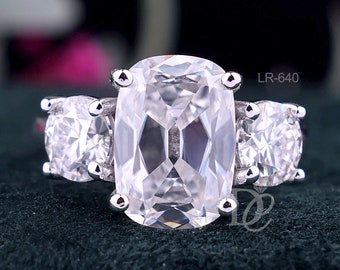 Puede incluir: Un anillo de tono plateado con una gran piedra central transparente de corte cojín, flanqueada por dos piedras transparentes más pequeñas de corte redondo. La banda de metal es pulida y reflectante. El texto LR-640 es visible arriba a la derecha.