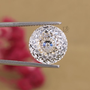 Può includere: Una pietra preziosa rotonda, trasparente e sfaccettata, tenuta da una pinzetta in metallo color argento. La pietra presenta un taglio complesso a molteplici sfaccettature che crea una superficie brillante e riflettente con un punto centrale.