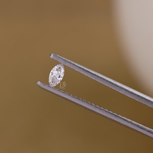 May include: A small, clear, marquise-cut gemstone held by a pair of silver-toned metal tweezers. The stone features a faceted surface that reflects light, and a small logo is visible on the surface of the stone near the center point.